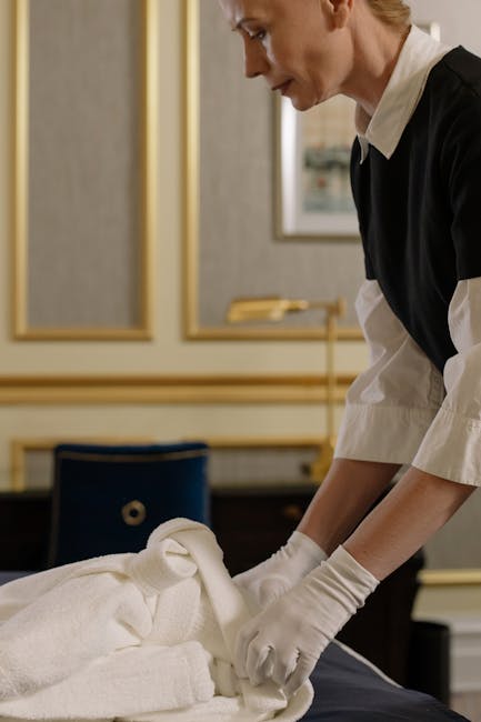 A professional cleaning technician from Deep Cleaning Knightsbridge, dressed in a black and white uniform, is carefully folding white towels on a table in a well-lit, elegant room with beige wallpaper, gold picture frame borders, and a dark wooden surface. The scene emphasizes surface cleaning and organisation within a domestic setting, showcasing attention to hygiene and meticulous care. The tidy arrangement and pristine appearance of the towels highlight the company's expertise in deep cleaning and sanitisation services suitable for residential spaces, aligning with the focus on avoiding hidden cleaning costs associated with overlooked cleaning tasks.