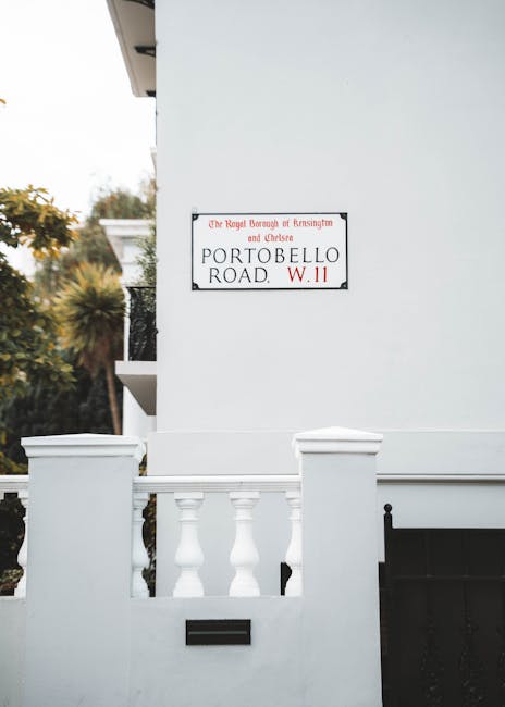 Close-up view of a white residential building corner with a white balustrade featuring decorative columns and a small black mailbox beneath a street sign. The sign, attached to the smooth, clean wall, indicates the location as Portobello Road, under the jurisdiction of Kensington and Chelsea council. Natural daylight illuminates the scene, highlighting the pristine surfaces and the contrast between the white exterior and the dark mailbox. In the background, blurred greenery and part of the building structure are visible. Deep Cleaning Knightsbridge offers professional surface cleaning and sanitisation services to maintain the cleanliness and hygiene of such residential exteriors, adhering to local council standards for waste disposal and environmental safety.