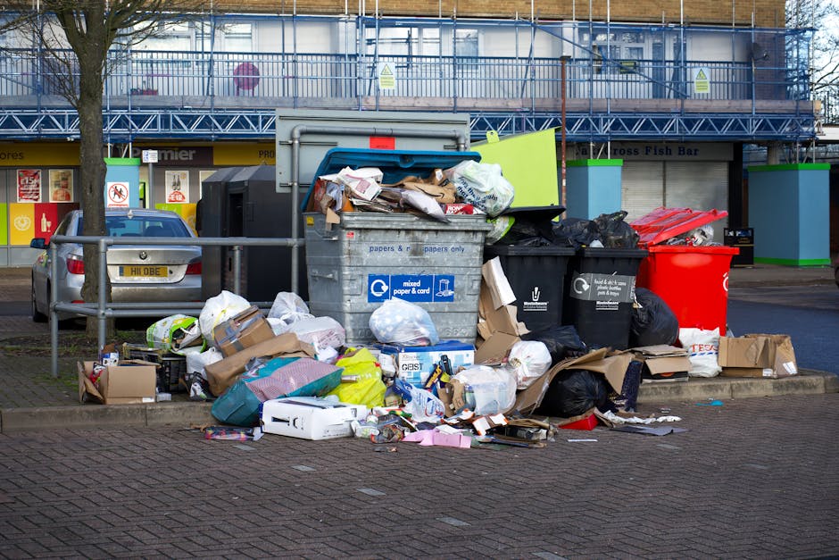 A large pile of mixed waste including cardboard boxes, plastic bags, paper, and household items is overflowing from a grey recycling bin labeled for mixed paper and card in an urban outdoor setting. Surrounding the bin, there are additional black and red waste containers, some filled with rubbish. The background shows a building with scaffolding and a blue railing, alongside a parked car. The pavement is partially covered with scattered trash, indicating improper waste disposal. Natural lighting highlights the cluttered scene, emphasizing the importance of proper waste management and hygiene in accordance with Kensington and Chelsea council rules for cleaning disposal, as promoted by Deep Cleaning Knightsbridge.