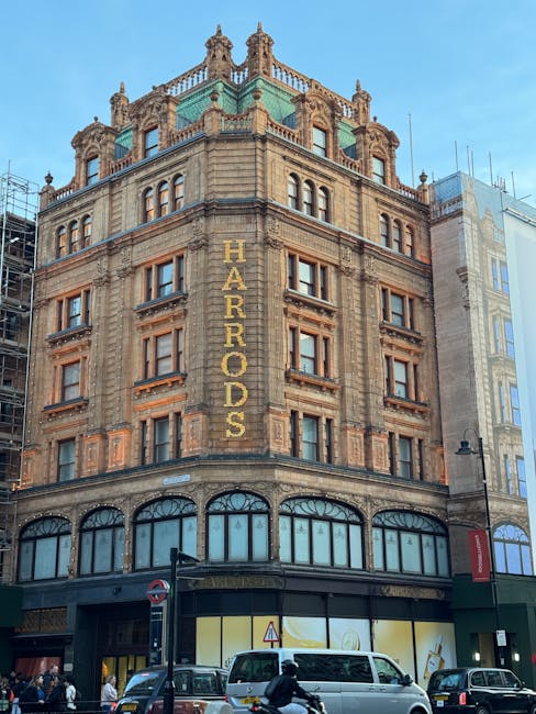 The image depicts a historic multi-story building with intricate architectural detailing and a stone facade, located in Knightsbridge near Harrods. The building features large arched ground-floor windows, some covered with reflective glass, and an ornate rooftop with decorative elements. The facade appears clean and well-maintained, indicative of thorough surface cleaning and preservation efforts. Adjacent modern buildings are visible to the right. The street level shows vehicles and pedestrians, with a lamppost and signage nearby. As experts in domestic and commercial cleaning, Deep Cleaning Knightsbridge specializes in surface cleaning, sanitisation, and deep cleaning services to maintain such architectural beauty.