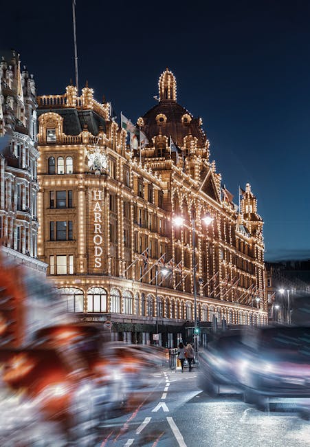 Nighttime view of the Harrods department store in Knightsbridge, illuminated with warm white lights outlining the building's ornate architectural details, including domes and towers. The street in front shows blurred motion of passing vehicles and pedestrians under streetlights, highlighting a lively urban atmosphere. The scene emphasizes the grandeur and elegance of the historic building, showcasing its intricate design and bright lighting against a dark sky. Image reflects the vibrant commercial environment that Deep Cleaning Knightsbridge may service for maintaining the pristine appearance of high-end retail spaces.