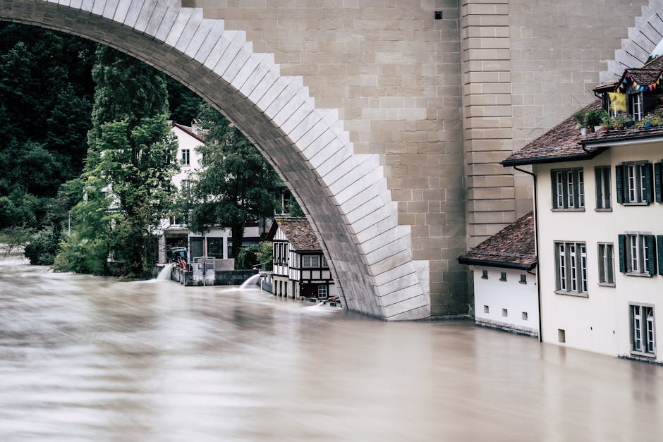 Image of a large stone bridge arching over a water-filled area, likely a river or floodplain, with surrounding greenery and residential buildings visible beneath and beside the bridge. The water appears brown and turbulent, indicating recent flooding. In the background, trees and part of a white house are visible, with some of the house's windows and rooftops partially submerged or close to the water level. The scene is well-lit with natural daylight, emphasizing the clean, structured surfaces of the bridge’s stonework as well as the wet environment. This setting highlights the importance of surface cleaning, sanitisation, and maintenance in flood-affected areas, as managed by Deep Cleaning Knightsbridge.