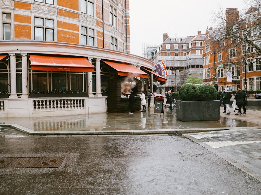 Street view in Knightsbridge showing a corner building with large windows, white decorative balustrades, and red awnings. The sidewalk appears wet from recent rain, reflecting the surroundings. Several pedestrians are walking and standing near a small outdoor seating area, with some wearing coats. A blackboard sign is placed outside a storefront, and a British flag is visible on a pole attached to the building. The area is lined with trees and nearby residential and commercial buildings, with scaffolding visible on one structure under renovation. The scene depicts an urban environment with clean, well-maintained surfaces and organized pedestrian space, consistent with professional deep cleaning and maintenance standards, as promoted by Deep Cleaning Knightsbridge.